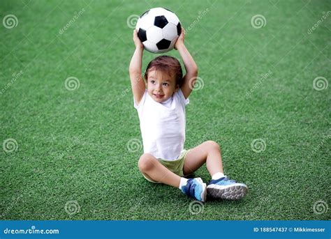 Toddler Boy In Sports Uniform Sitting On Green Footall Field Holding