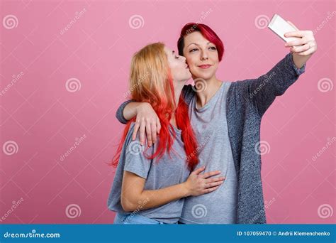 two lesbian girls embrace and kiss on the cheek and do selfie on a mobile phone on a pink