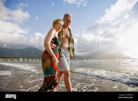 Happy Heterosexual Couple Laugh As They Walk With Their Arms Around Each Other On A Beach At