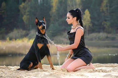 An Attractive Girl In A Black Dress With A Doberman Sitting On The Sand