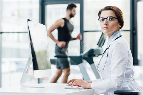Doctor In Glasses Using Computer While Sportsman Running On Treadmill