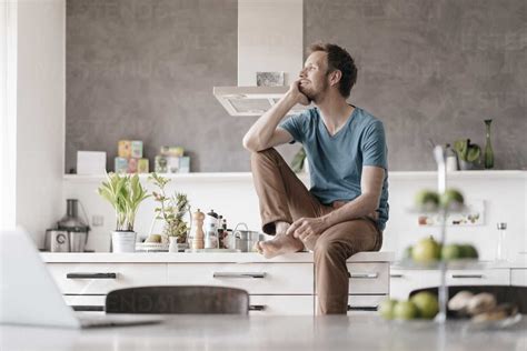 Smilingg Man Sitting On Kitchen Counter Looking At Distance Stock Photo