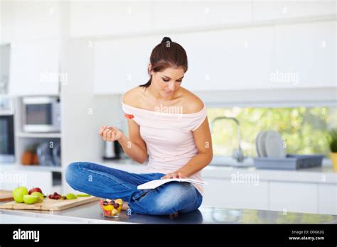 An Attractive Young Woman Sitting On The Kitchen Counter Reading A And