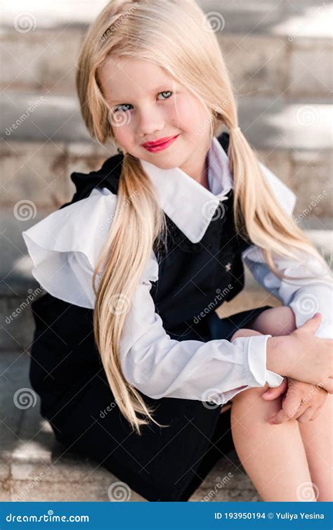 Маленькая белокурая девушка с 2 кабелями в школьной форме сидит в улыбках и представлениях шагов