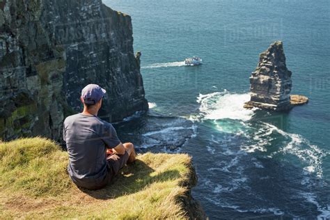 Man Sitting On Grassy Cliffs Edge Overlooking Large Sea Stack Rock