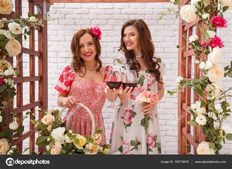 Two Beautiful Girls Dressed In Summer Dresses Posing Near A Flower Arch With Glasses Of Red Wine