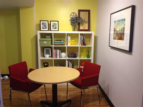 Two Red Chairs Sit At A Round Table In Front Of A Bookcase With Books On It