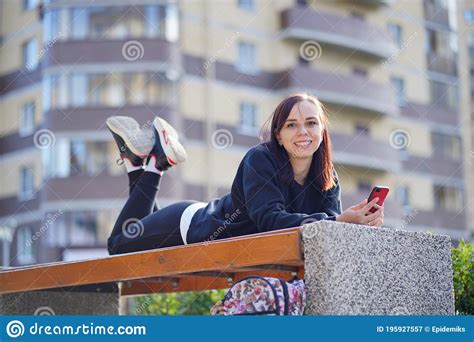 Young Woman Lying On Her Stomach Outside On A Bench With Her Legs Up And Browsing Her Cell