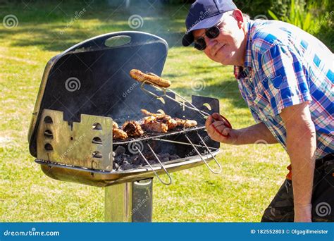 A Man At The Charcoal Grill Is Holding A Grilled Sausage With The Tongs In His Hand And Is
