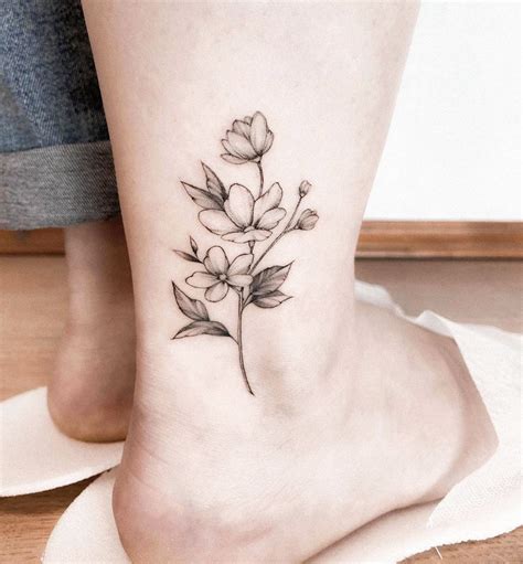 A Small Flower Tattoo On The Ankle Is Shown In Black And Grey Ink With Leaves Coming Out Of It