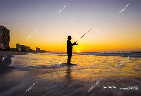 Blurred View Of Silhouette Of Man Fishing In Waves On Beach At Sunset