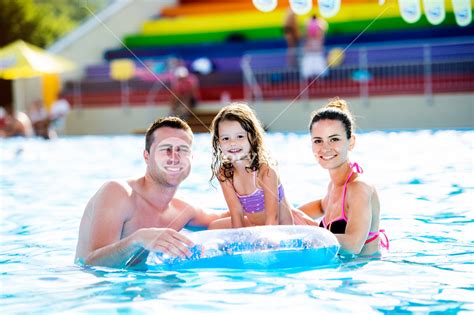 Young Mother And Father With Their Daughter Sitting On Pool Lilo In Swimming Pool In Aqua Park
