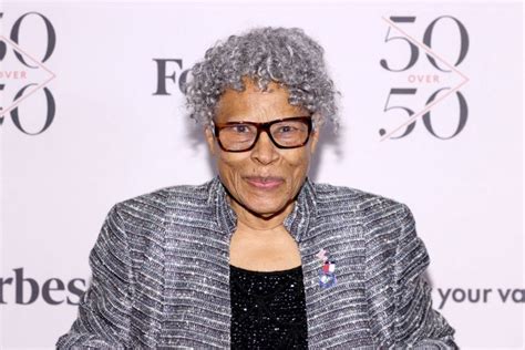 An Older Woman Wearing Glasses Standing In Front Of A White Background