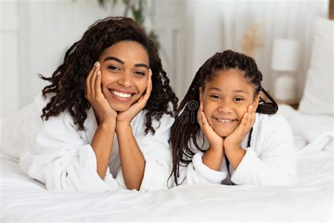 Portrait Of Happy Black Mother And Daughter In Bathrobes Posing On Bed
