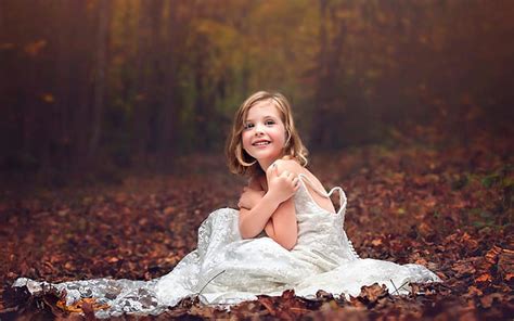 स्माइली प्यारा छोटी लड़की वन पृष्ठभूमि में सूखी पत्तियों पर बैठे सफेद