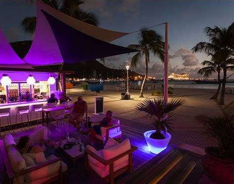 People Sitting At Tables On The Beach In Front Of An Umbrella Covered Bar With Purple Lights