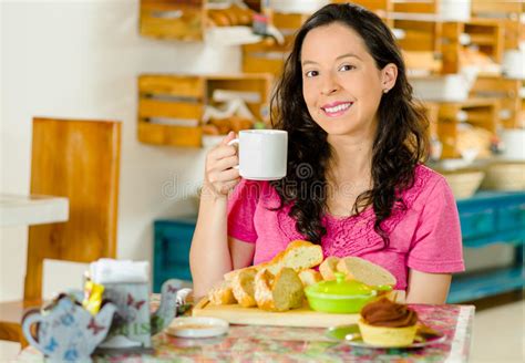 pretty brunette woman sitting at table inside bakery holding cup of coffee and smiling happily