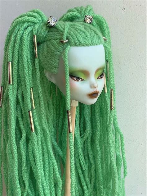 A Doll With Green Dreadlocks Is Wearing A Wig And Holding A Wooden Stick