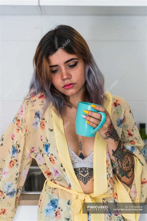 mujer joven y somnolienta en bata de seda sosteniendo taza de bebida caliente mientras está de