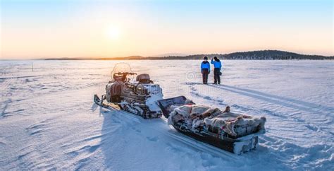 Young Couple Enjoy Sunrise Near Snowmobile On Frozen Lake Inari In