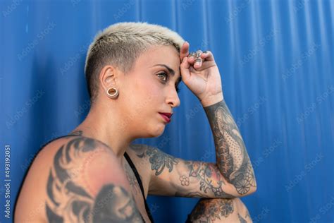 atractiva mujer con cuerpo tatuado y pelo rubio muy corto sobre un fondo azul y blanco stock