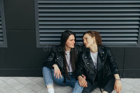 Close Up Photo Of Two Smiling Girls In Leather Jackets And Jeans Sitting On A Cobblestone