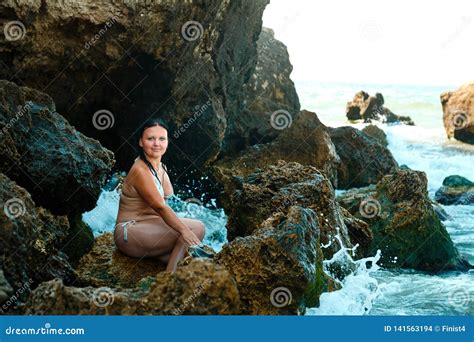 A Young Brunette In A White Bathing Suit Is Sitting On A Rock At The