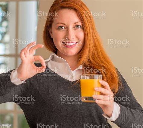Redhead Woman Holding Orange Juice Glass At Home Doing Ok Sign With