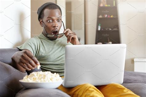 Surprised African Male Sitting On Couch At Home Eating Popcorn And Watching Exciting Tv Show