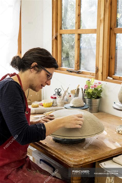 Woman In Red Apron Sitting In Ceramics Workshop Working On Clay Bowl