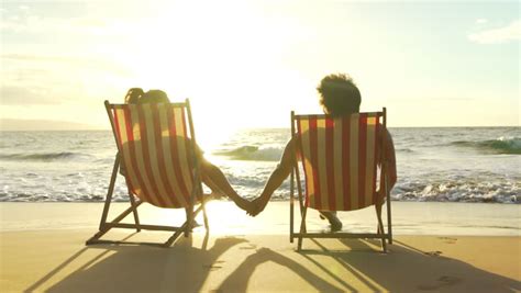 Romantic Couple Sitting In Wooden Deckchairs On The Beach Toasting The