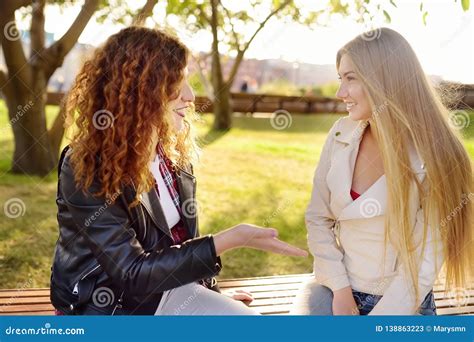 Two Beautiful Young Women Talking While Sitting On A Bench At Sunny