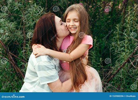Young Mom Kisses Her Little Daughter On The Cheek Against The Green