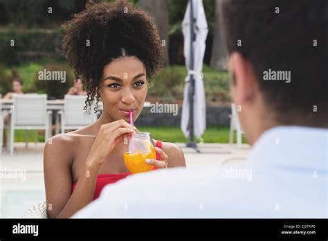 Romantic Couple Of Multiethnic Young People Sitting By The Swimming Pool Drinking Juicy Orange
