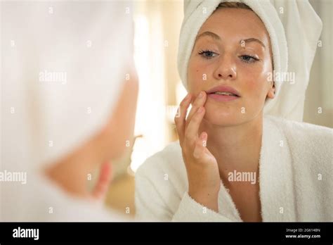 Caucasian Woman In Bathroom Wearing Bathrobe And Towel Looking In Mirror And Moisturising Face