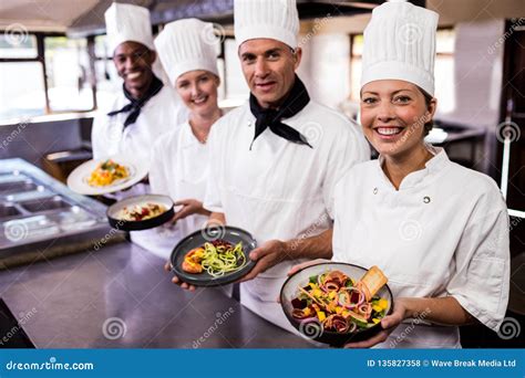 Group Of Chefs Holding Plate Of Prepared Food In Kitchen Stock Photo