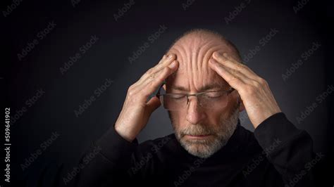 Tired 60 Years Old Man With A Beard And Glasses Massaging His Forehead A Headshot Against A