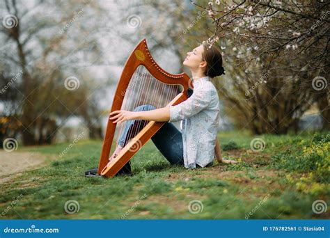 Woman Harpist Sits On Grass And Plays Harp Among Blooming Apricot Trees
