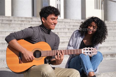 A Young Latin Couple Playing The Guitar And Smiling Sitting On Stairs