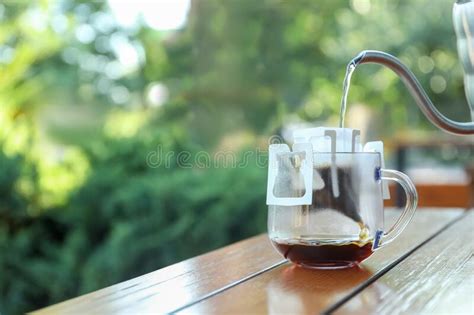 Pouring Hot Water Into Glass Cup With Drip Coffee Bag From Kettle On Wooden Table Outdoors