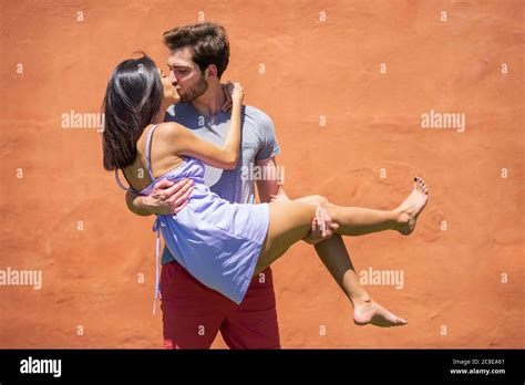Young Man Kissing While Carrying Woman Against Brown Wall At Back Yard During Sunny Day Stock