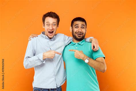 two best friends hugging pointing finger each other and looking at camera with shocked face