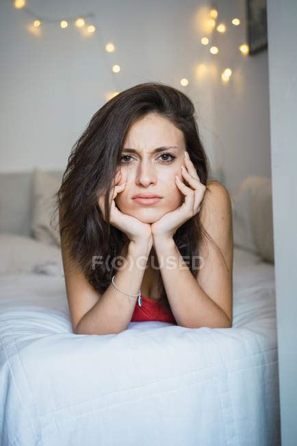 displeased brunette girl lying on her bed with head on her hands and looking at camera
