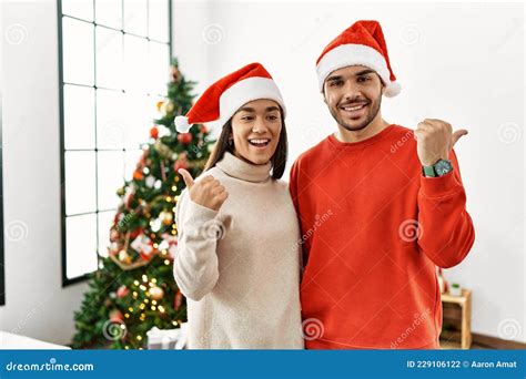 Young Hispanic Couple Standing By Christmas Tree Smiling With Happy