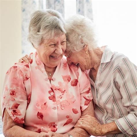 Two Older Women Laugh Together While Sitting On A Bed Women Laughing