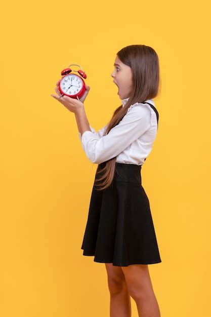 Шокированная девочка подросток в школьной форме с будильником показывающим время на желтом фоне