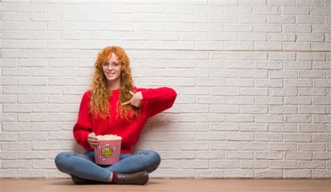 Young Redhead Woman Sitting Over Brick Wall Eating Popcorn With Surprise Face Pointing Finger To