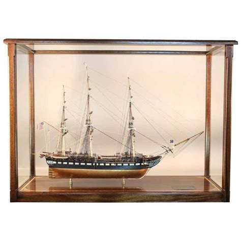 A Model Ship In A Glass Case On A Wooden Shelf With White Walls Behind It