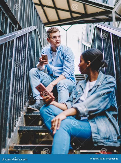 Multicultural Young Man And Woman With Mobile Phones Sitting On Stairs