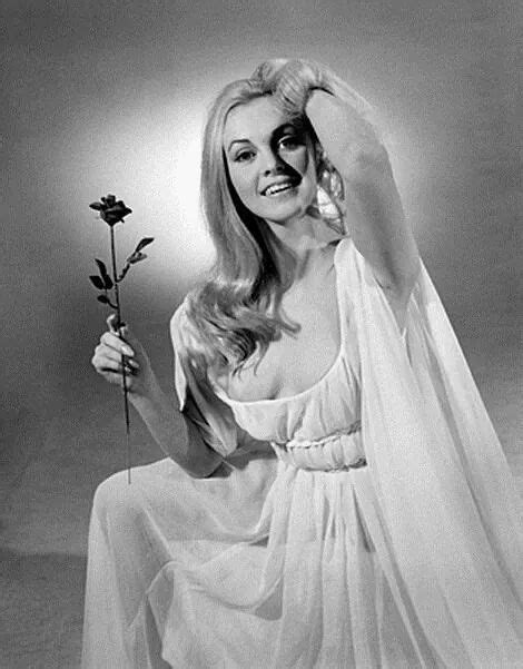 A Woman In A White Dress Holding A Flower And Posing For The Camera With Her Hands Behind Her Head